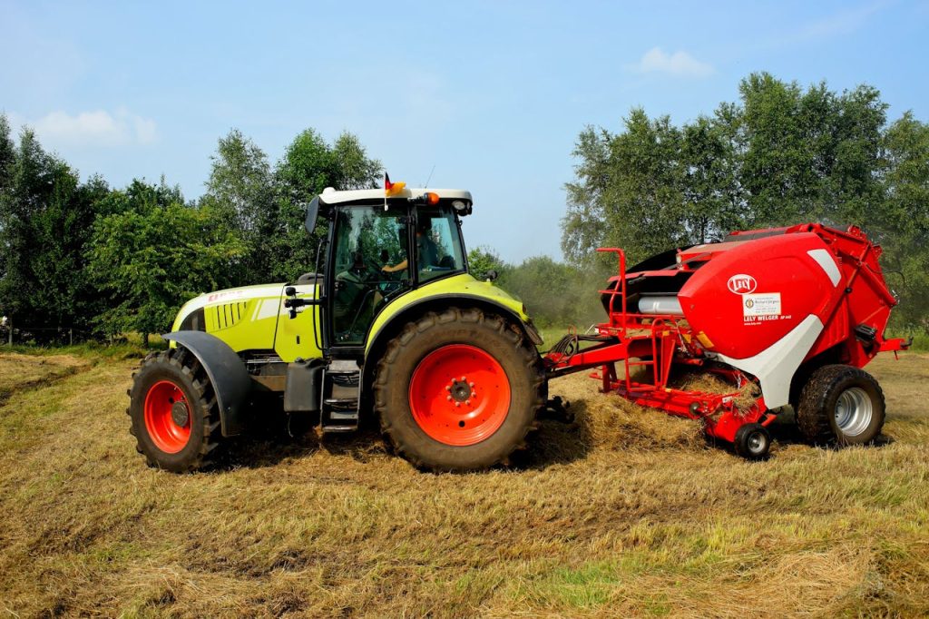 Green tractor and hay baler working on a farm field with trees in the background.